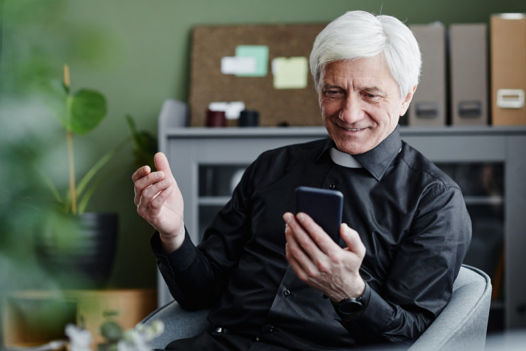 Fotografia di un prete che guarda sorridente lo schermo del proprio smartphone
