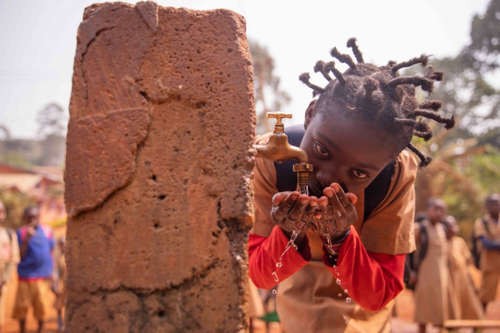 Fotografia di una bambina che si abbevera con le mani in una fontanella in un villaggio africano
