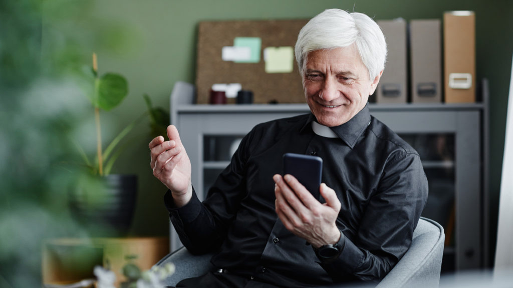 Fotografia di un prete che guarda sorridente lo schermo del proprio smartphone